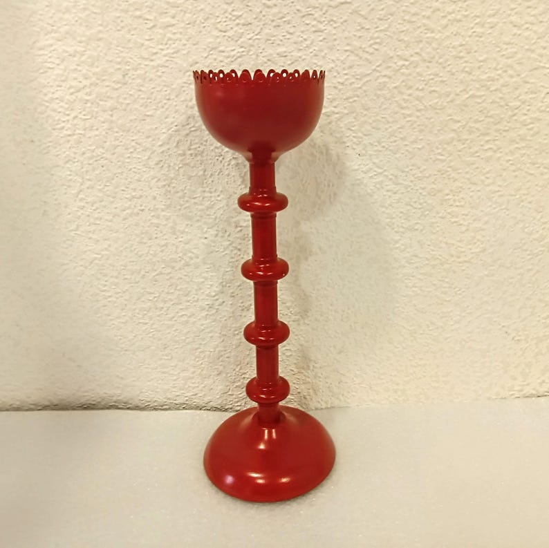 May include: A bright red metal candle holder featuring a tall, slender stem with three distinct horizontal ring accents. The top section is a bowl-shaped cup with a scalloped, decorative edge, and the base is a wide, circular, flat disc.