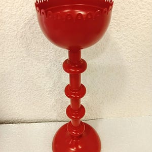 May include: A bright red metal candle holder featuring a tall, slender stem with three distinct circular rings and a wide, stable base. The top section is a deep, bowl-shaped cup with a decorative scalloped edge along the upper rim.