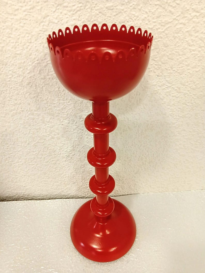 May include: A bright red metal candle holder featuring a tall, slender stem with three distinct circular rings and a wide, stable base. The top section is a deep, bowl-shaped cup with a decorative scalloped edge along the upper rim.