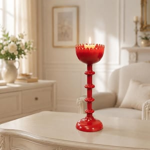 May include: A tall, bright red candle holder with a scalloped, bowl-shaped top and a slender, multi-tiered stem. A single, lit cream-colored pillar candle sits securely within the bowl, casting a warm glow from the center of the red structure.