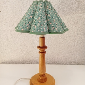 Puede incluir: Una lámpara de mesa vintage con base de madera y pantalla floral verde. La pantalla presenta un patrón de pequeñas flores rosas sobre fondo verde. La lámpara tiene un diseño clásico, ideal para un dormitorio o salón.