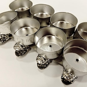 Puede incluir: Un conjunto de ocho pequeños moldes redondos de metal plateado, cada uno con un pequeño agujero en el centro. Los moldes están conectados por una base metálica decorativa y texturizada. Los moldes están dispuestos en un patrón circular.
