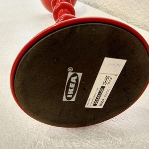 May include: The underside of a bright red object with a circular base. A black felt pad covers the bottom, featuring a white IKEA logo and a rectangular product label with the text 302.701.23, 20529, and Made in China printed on it.