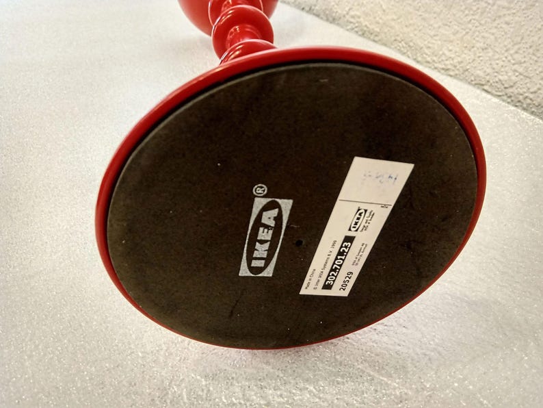 May include: The underside of a bright red object with a circular base. A black felt pad covers the bottom, featuring a white IKEA logo and a rectangular product label with the text 302.701.23, 20529, and Made in China printed on it.
