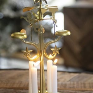 May include: A gold-colored Christmas candle carousel with a star on top and star-shaped accents. The carousel holds white candles that are lit, and the carousel is on a gold-colored tray.