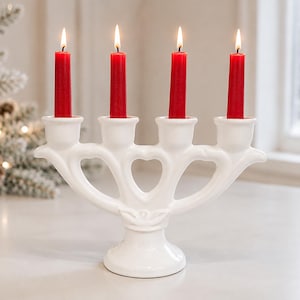 Puede incluir: Un candelabro de cerámica blanca con cuatro portavelas dispuestos en un diseño curvo y simétrico con recortes en forma de corazón. Cuatro velas cónicas rojas y delgadas están insertadas, cada una con una llama encendida en la parte superior.