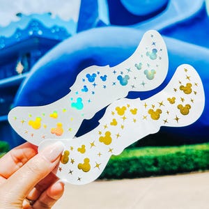 May include: Two clear stickers with Mickey Mouse shapes and stars in gold, blue, and iridescent hues. The stickers are held up against a blue sculpture and a bright sky, suggesting a theme park setting.