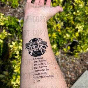 K&ouml;nnte beinhalten: Ein schwarzes und wei&szlig;es tempor&auml;res Tattoo mit dem Universal Studios Logo und einer Liste von Restaurants und Bars, darunter Finnegan's, Chez Alcatraz, The Hopping Pot, Duff Brewery, Backwater Bar, Hogs Head, The Watering Hole und Cart.