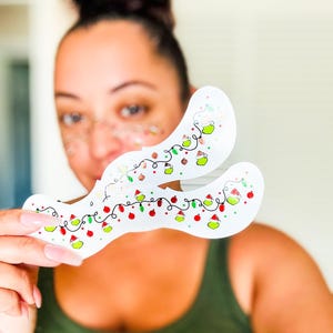 May include: Two white, curved eye mask stencils decorated with a string of Christmas lights, red and green ornaments, and small green trees. The stencils are held up in front of a person.