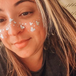 May include: A person with silver Mickey Mouse and star face stickers. The person has long, light brown hair with blonde highlights and is wearing a dark grey top. The person is smiling.