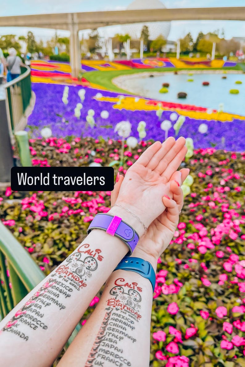 May include: Two people's arms with temporary tattoos that list countries they have visited. The tattoos say "Cali's Birthday" and list countries like Norway, Mexico, China, Germany, Italy, USA, Japan, Morocco, France, and Canada. The tattoos are on their forearms and they are holding their hands up in front of a colorful flower bed.