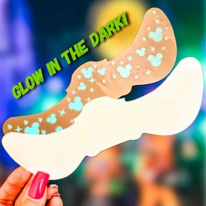 May include: Two beige, curved adhesive products, one with light blue Mickey Mouse head and star patterns, and the other plain white. The text "GLOW IN THE DARK!" is in green. The products are held by a hand with a pink nail.