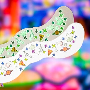 Op de afbeelding: Twee vellen met golvende randen voorzien van een herhalend patroon van ruimte-thema iconen, waaronder groene alienhoofden, planeten met ringen, pizzapunten, grijparmen en vierpuntige sterren in diverse kleuren op een witte en lichtgrijze achtergrond.