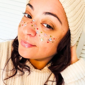 May include: A person wearing a cream-coloured knitted beanie and a matching sweater. The person's face is decorated with festive, colourful temporary tattoos, including snowflakes, hearts, and candy canes, creating a holiday-themed look.