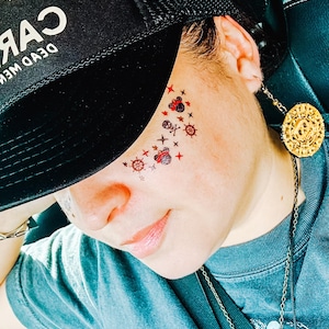 May include: A black trucker hat with the text "DEAD MEN TELL NO TALES" is worn. Temporary tattoos of red stars, skulls, and ship wheels are on the face. A gold coin pendant hangs from the ear.