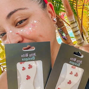 May include: Two packages of white, decorative face stickers with red and white swirl designs. The packages are held up in front of a person's face. The packages have the text "little empires" printed on them.