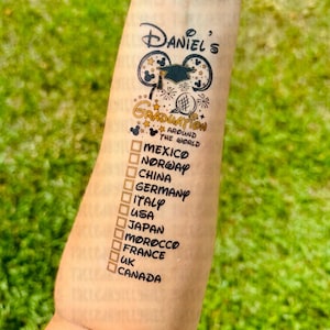 May include: A temporary tattoo with a graduation theme. It features a Mickey Mouse ear design with a graduation cap and the text "Daniel's Graduation Around the World". Below the design is a list of countries with checkboxes: Mexico, Norway, China, Germany, Italy, USA, Japan, Morocco, France, UK, Canada.