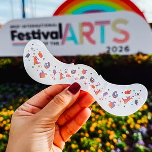 May include: A white, mustache-shaped sticker with colourful cartoon illustrations. The sticker is held in front of a sign that reads "Festival of the ARTS 2026" with a rainbow in the background. The sticker is decorated with various cartoon characters.