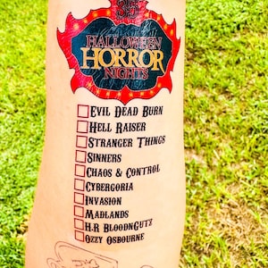 Puede incluir: Un tatuaje temporal con el logotipo rojo y negro de Halloween Horror Nights 35 sobre una lista de diez elementos: Evil Dead Burn, Hell Raiser, Stranger Things, Sinners, Chaos & Control, Cybergoria, Invasion, Madlands, H.R. Bloodngutz y Ozzy Osbourne.