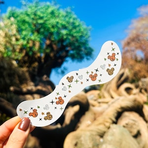 Puede incluir: Una pegatina blanca y ondulada con cabezas de Mickey Mouse con rayas de tigre y estampado de leopardo, junto con estrellas y cruces. La pegatina se sostiene contra un fondo de cielo azul y exuberante vegetación.