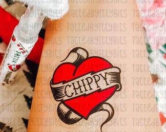 May include: A temporary tattoo featuring a red heart with a banner that reads "CHIPPY" in a classic tattoo style. The tattoo is applied to the skin, with a partially visible applicator in the background.
