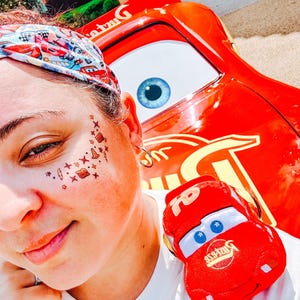 May include: A person wearing a white t-shirt with a Cars graphic, a Cars-themed headband, and temporary face tattoos. A plush Lightning McQueen car is on the person's shoulder. A large red Lightning McQueen car is in the background.