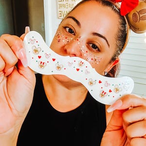 Puede incluir: Una mascarilla facial blanca y curvada decorada con cabezas de Mickey Mouse, corazones y mariposas. La mascarilla se sostiene frente a la cara de una persona. La persona lleva una camiseta negra y una diadema de orejas de jengibre de Mickey Mouse.