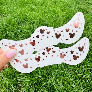 May include: Two white, curved decorative pieces with a pattern of copper-colored Mickey Mouse heads and stars. The pieces are held against a background of green grass.