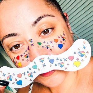 May include: A face decorated with heart and star stickers in various colours. A white, mustache-shaped prop is held up, decorated with hearts and stars, including rainbow patterns.