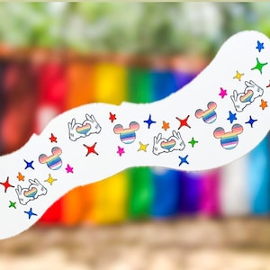 May include: A white, curved decorative piece with colourful Mickey Mouse head designs, hand gestures, and star shapes. The designs incorporate rainbow colours, symbolising LGBTQ+ pride. The background is a blurred rainbow gradient.