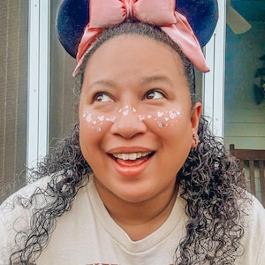 Puede incluir: Una diadema con orejas de Minnie Mouse negras y rosas con un gran lazo rosa. La persona lleva una camiseta beige con las palabras "Mickey & Minnie's" impresas. La persona tiene purpurina en la cara.