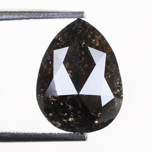 Może przedstawiać: Czarny diament w kształcie gruszki o szlifie fasetowym i ciemnoszarym kolorze. Diament jest trzymany przez pęsetę.