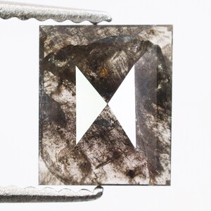 May include: A square-cut, salt and pepper diamond held by tweezers. The diamond features a unique pattern of black and gray inclusions, with a white, geometric design in the center. The stone is set against a white background.