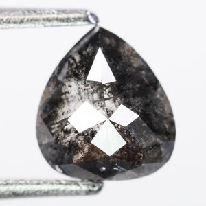 Op de afbeelding: Een peervormige zwarte diamant met een facetgeslepen vorm en insluitsels. De diamant wordt vastgehouden door een pincet tegen een witte achtergrond.