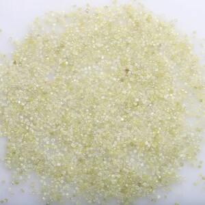 Può includere: Un'immagine ravvicinata di un mucchio di piccoli diamanti sfaccettati di colore giallo chiaro. I diamanti sono sparsi su una superficie bianca.