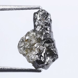Op de afbeelding: Een ruwe, donkergrijze diamant met een zilveren glans, vastgehouden door een pincet tegen een witte achtergrond.