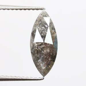 Puede incluir: Un diamante de talla marquesa sujeto con pinzas. El diamante tiene una mezcla de facetas grises y claras, con forma puntiaguda. Las facetas reflejan la luz, creando un efecto brillante. El fondo es blanco liso.