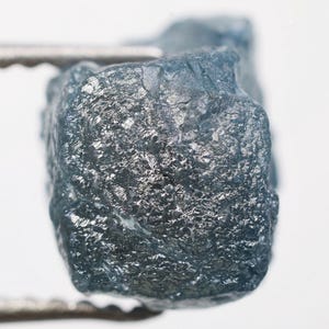 May include: A raw, unpolished, blue-grey gemstone held by tweezers. The surface of the stone has a textured, crystalline appearance. The background is plain white, highlighting the natural beauty of the mineral specimen.