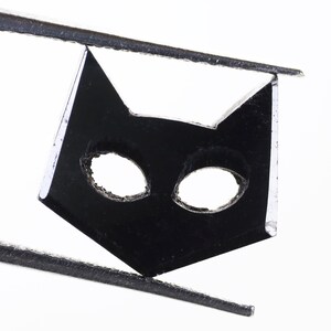 May include: A black cat-shaped cutout with two eye holes, held by tweezers against a white background.