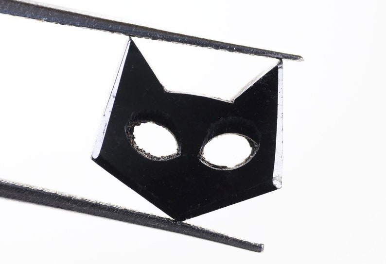 May include: A black cat-shaped cutout with two eye holes, held by tweezers against a white background.