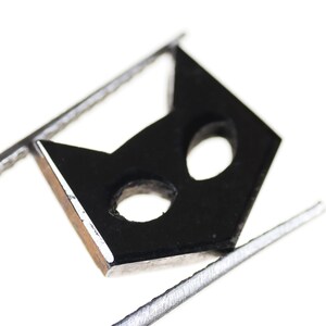 May include: A black cat mask cutout with two eye holes, held by silver tweezers against a white background.