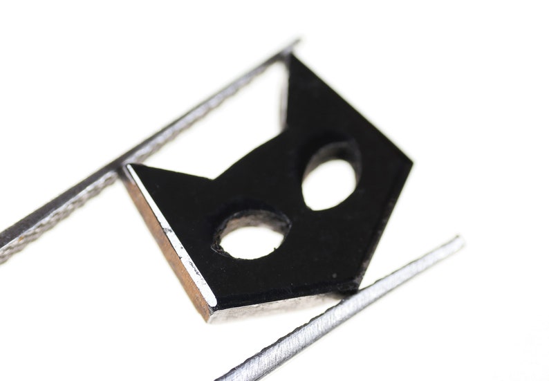 May include: A black cat mask cutout with two eye holes, held by silver tweezers against a white background.