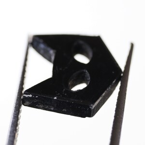 May include: A black plastic button with two holes in the shape of an arrow. The button is held in place by two silver tweezers.