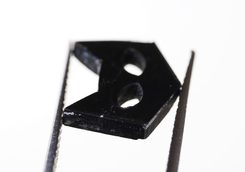 May include: A black plastic button with two holes in the shape of an arrow. The button is held in place by two silver tweezers.
