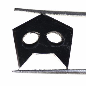 May include: A black metal object with two holes and a pointed tip, held by silver tweezers.