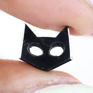 May include: A black cat-shaped charm with cut-out eyes, held between two fingers.