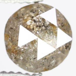 Op de afbeelding: Een ronde, ruwe diamant met een gespikkelde, lichtbruine en grijze uitstraling. De diamant heeft drie grote, witte, driehoekige facetten. De edelsteen wordt vastgehouden door een zilveren pincet tegen een witte achtergrond.