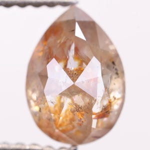 Puede incluir: Una gema facetada en forma de pera, con un tono ámbar cálido. La gema muestra inclusiones internas y un corte complejo y multifacético, creando un juego de luces y sombras. La piedra está sujeta por una pinza metálica.