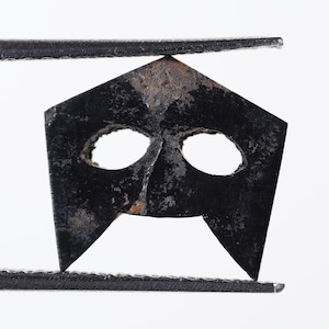 May include: A black metal mask with two eye holes and a pointed chin. The mask is held by tweezers.
