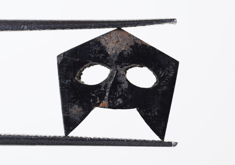May include: A black metal mask with two eye holes and a pointed chin. The mask is held by tweezers.